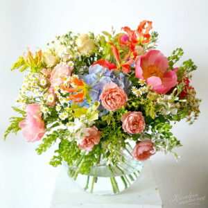 Wiosenny bukiet z różowymi różami, niebieskimi hortensjami, żółtymi kwiatami, oranżowymi gloriozami oraz białymi stokrotkami, ułożony w przezroczystym wazonie na białym tle.